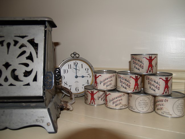 A photo of a countertop holding a toaster with a perforated metal design, a clock, and eight cans, labeled ‘Original Deviled Ham’ and ‘Underwood Co Boston Mass’ featuring a red devil standing with his arms out.