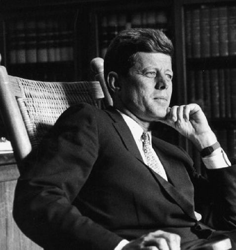 President Kennedy is seated sideways in a rocking chair looking ahead while resting his chin in his hand.  He looks like he's deep in thought.