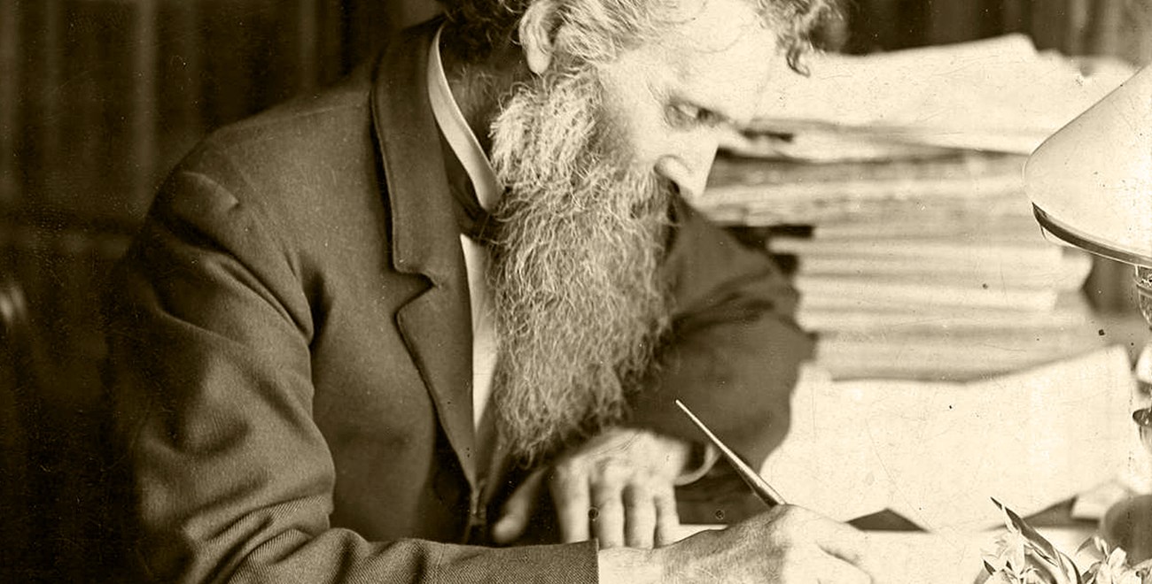 A mature John Muir, writing at his desk in suit and tie.