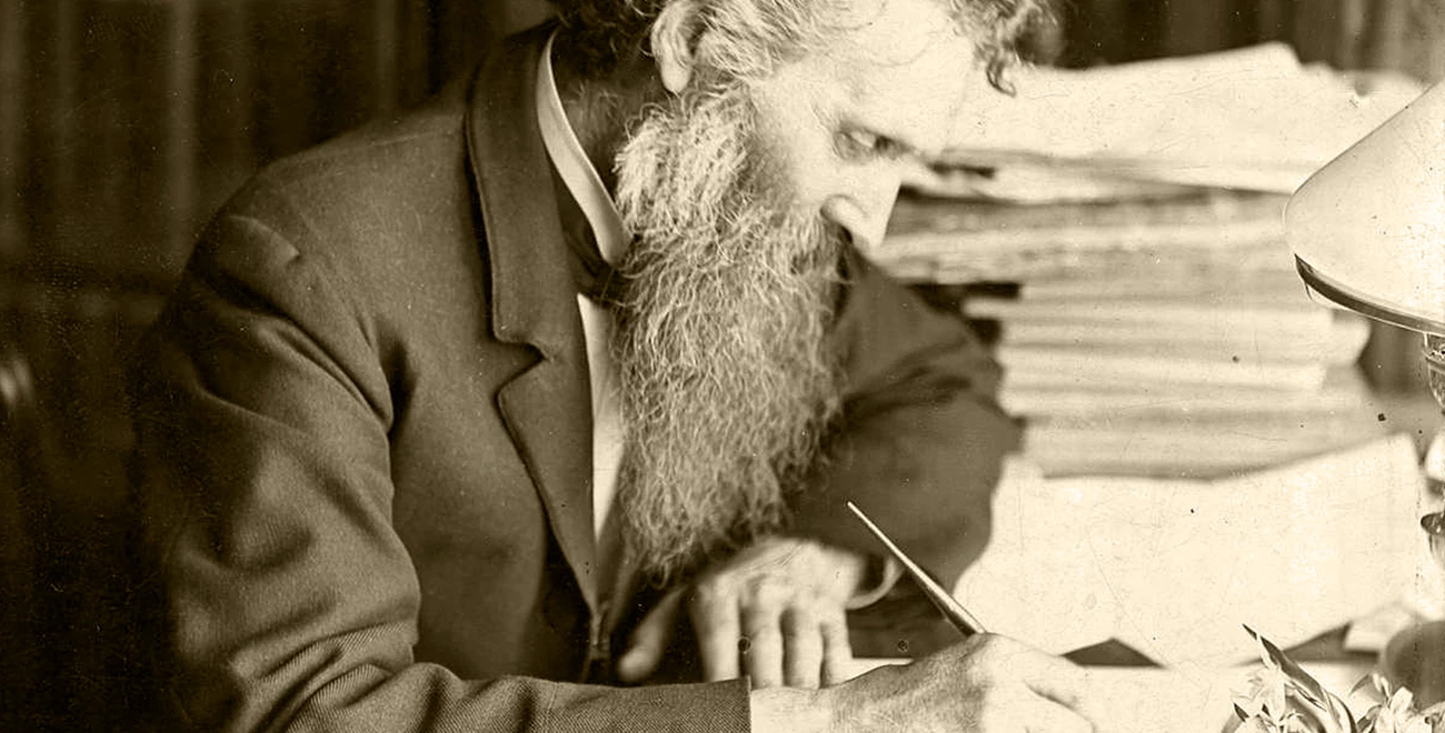 John Muir at his writing desk Historic photograph of John Muir seated at a desk, writing with a pen in hand. Papers and a lamp are visible on the desk, reflecting his dedication to documenting his thoughts and advocating for conservation.