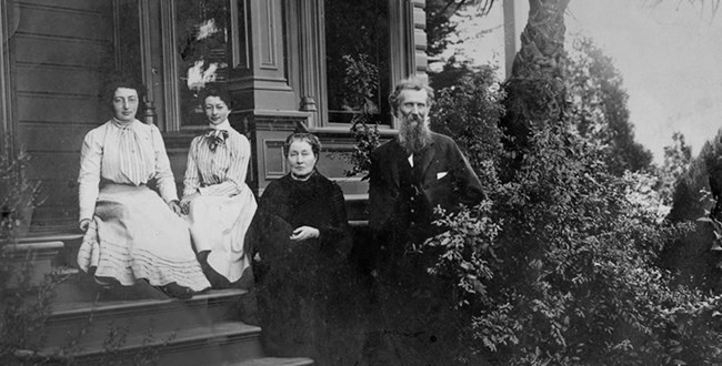 An older man in a suite with a beard, two younger women in dresses and an older woman in a dress sit on a porch, surrounded by manicured bushes.