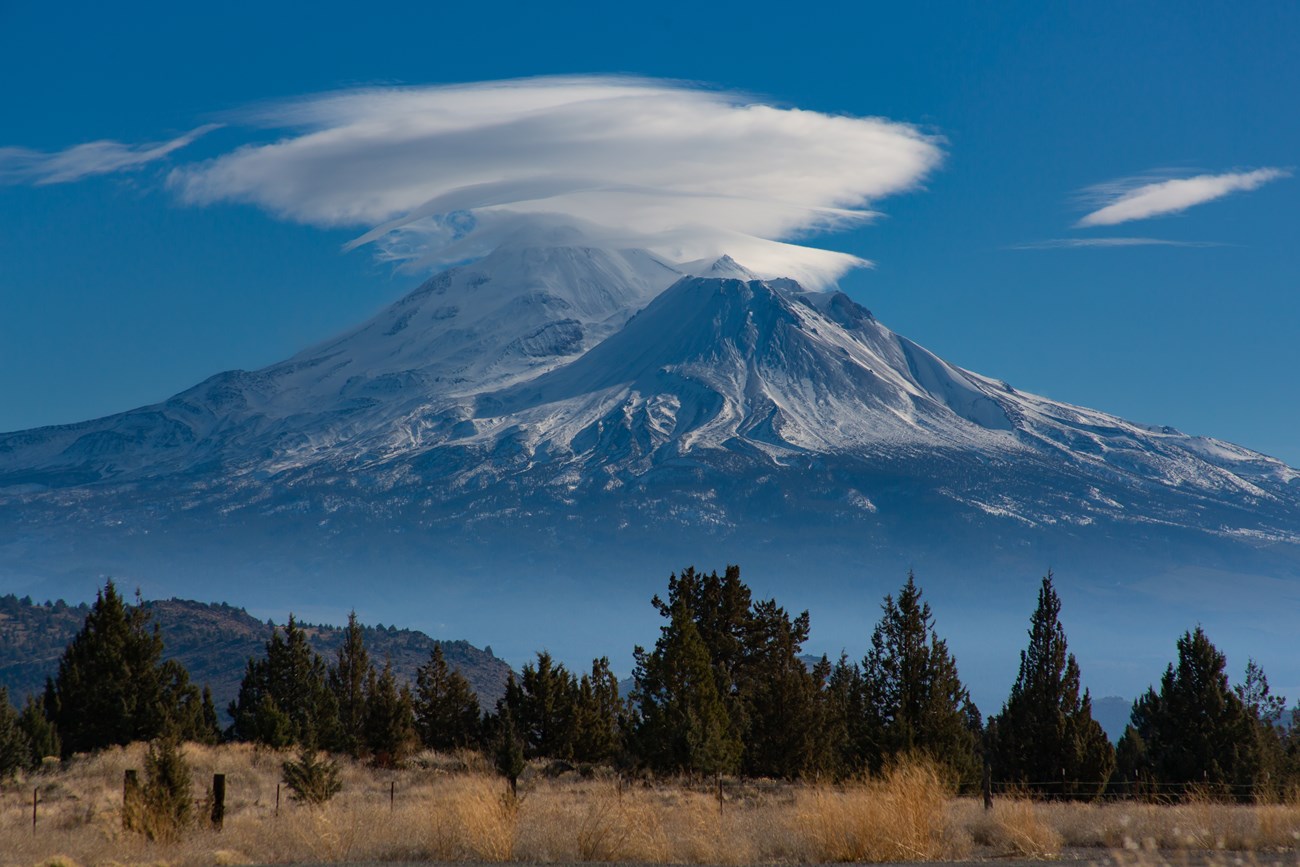 A majestic view of Mount Shasta with snow-covered peaks and a distinctive lenticular cloud hovering above. The scene includes a foreground of trees and dry grassland, set against a bright blue sky.