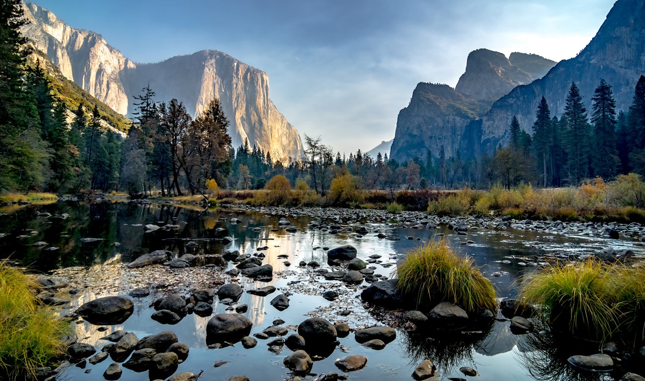 A scenic view of Yosemite National Park showcasing a tranquil river with smooth rocks, surrounded by lush green trees, and towering granite cliffs illuminated by the setting sun.