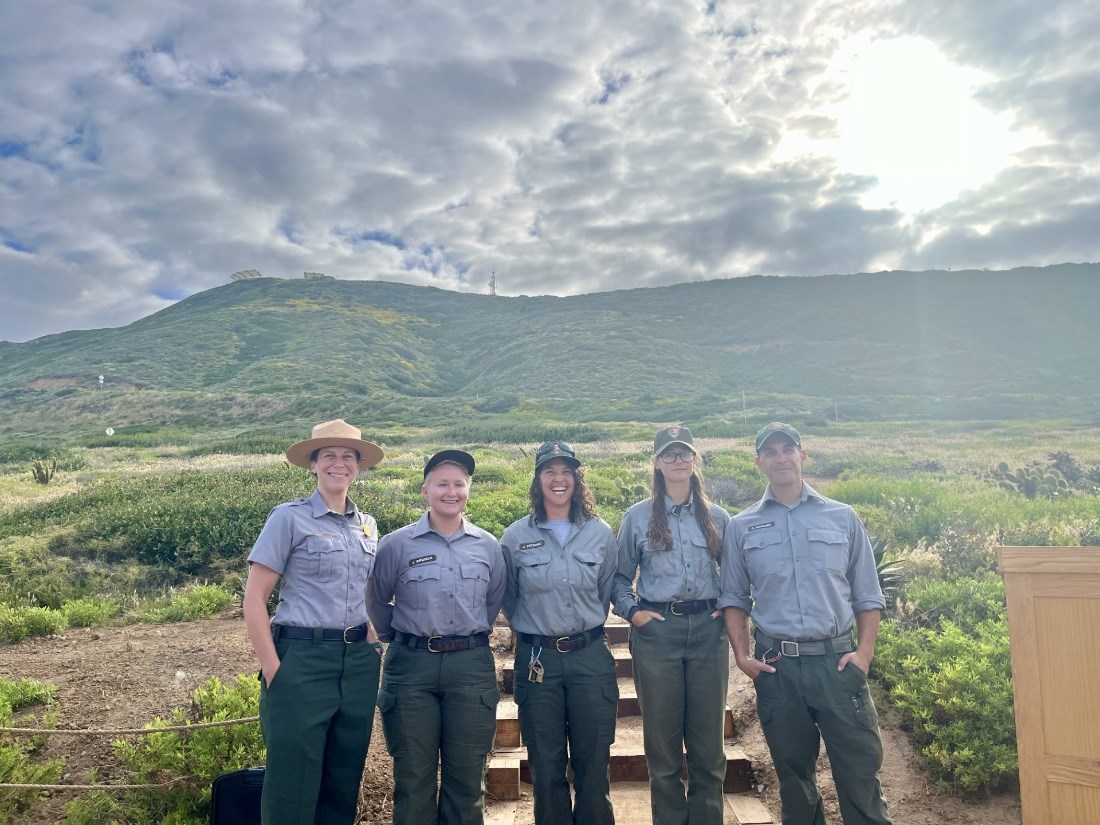 Five uniformed park rangers in front of large grassy hills.