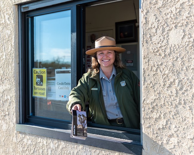Alt Text: A Park Ranger smiling and holding out a brochure from an entrance booth.