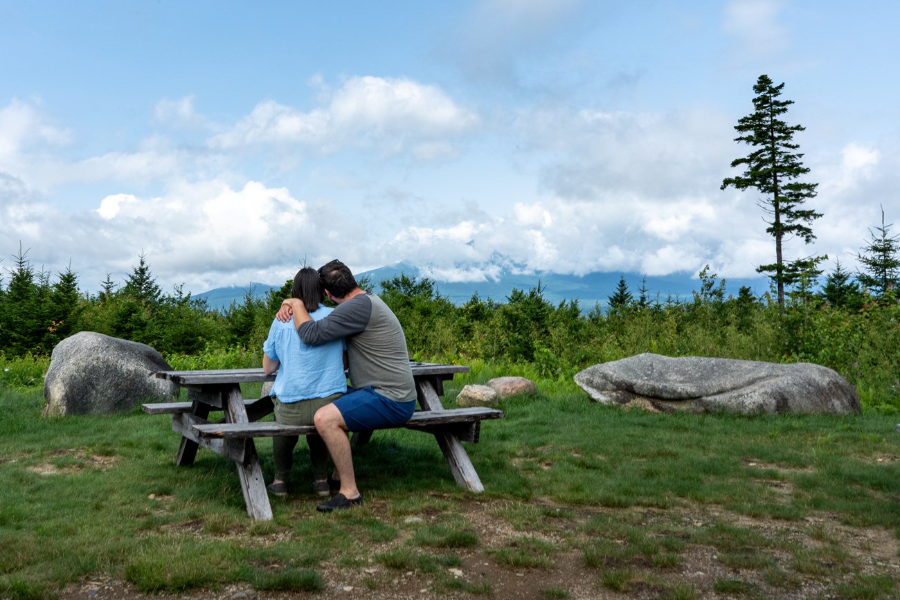 A woman and man sit at a picnic table on a grassy surface. They sit with their backs towards us and face a view of Mount Katahdin in the distance. It is a sunny blue sky day with patchy clouds in the sky.
