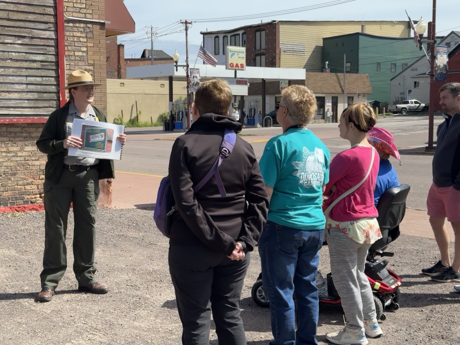 A park ranger holding a photograph stands in front of a group gathered on the sidewalk near a historic sign