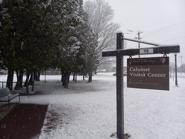 A blanket of white snow covers the ground and trees surrounding a sign that reads Calumet Visitor Center.