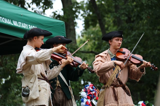 A man in 18th century clothing is demonstrating a drum to 3 guests.