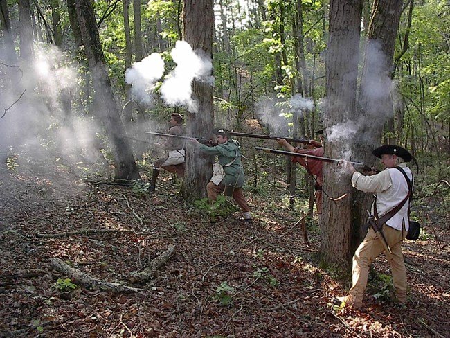 A line of soliders in 18th century uniforms are firing muskets towards the left.