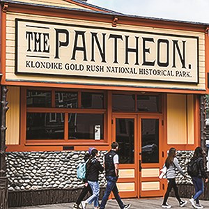 Pantheon Saloon is a yellow building with bright orange trim.  Pantheon Saloon is positioned across the top of the building in large black letters. A few people walk along the boardwalk in front of the building.