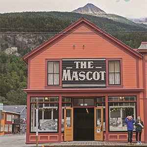Two people taking pictures in front of a burnt orange building titled "The Mascot" in large white letters with a mountain peak in the background.