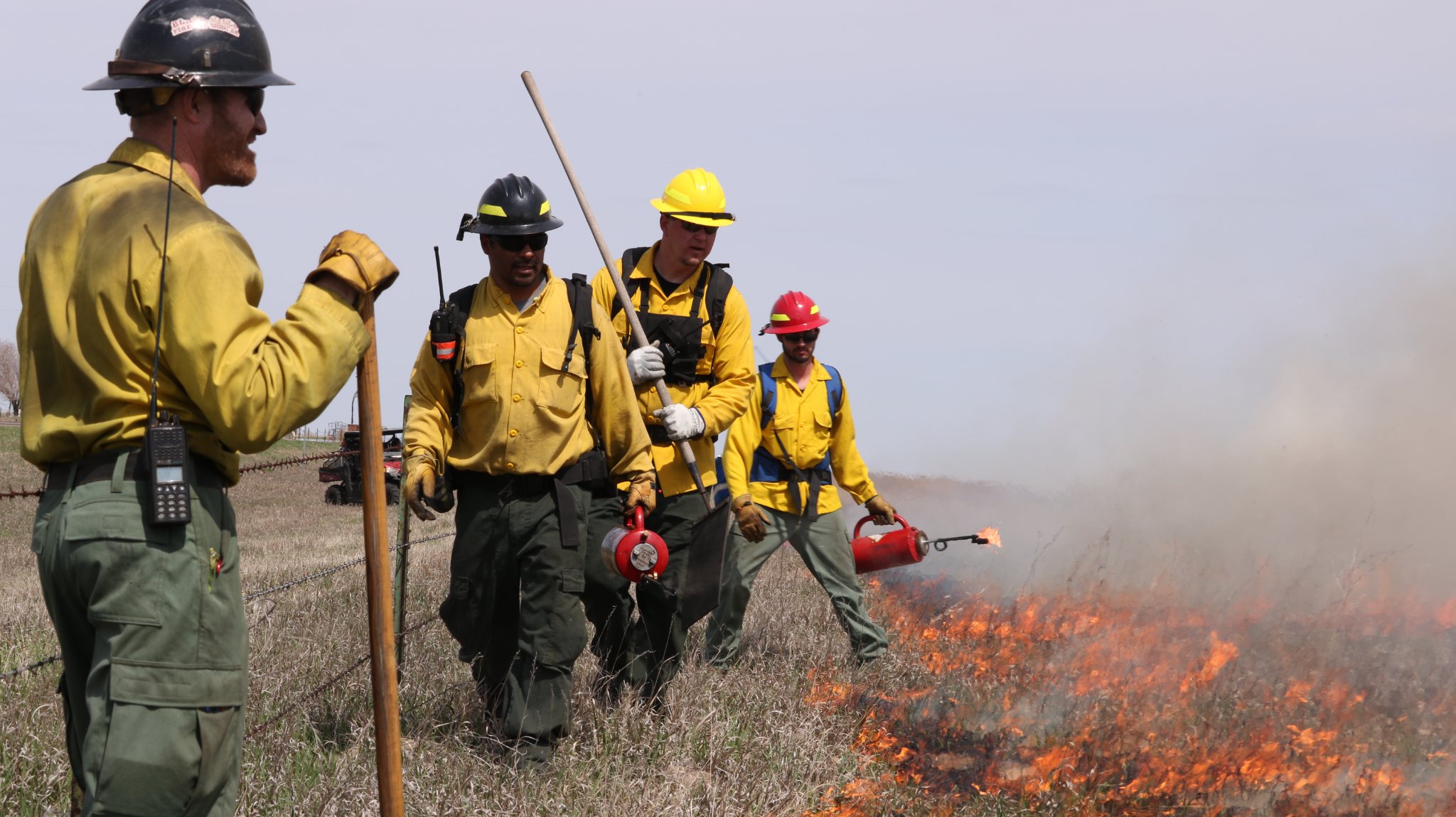 Firefighters stand along a line of fire with various tools.