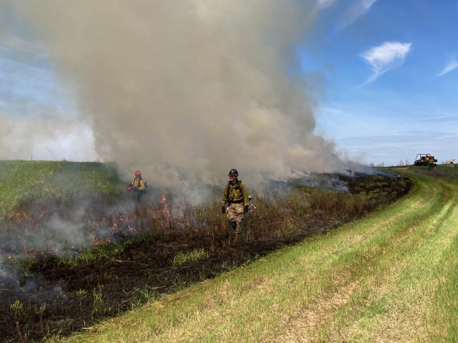 dan with gloves Fire fighters ignite fire while walking in the black of the burn. The grass is green and black. The sky is a vibrant blue with white clouds.