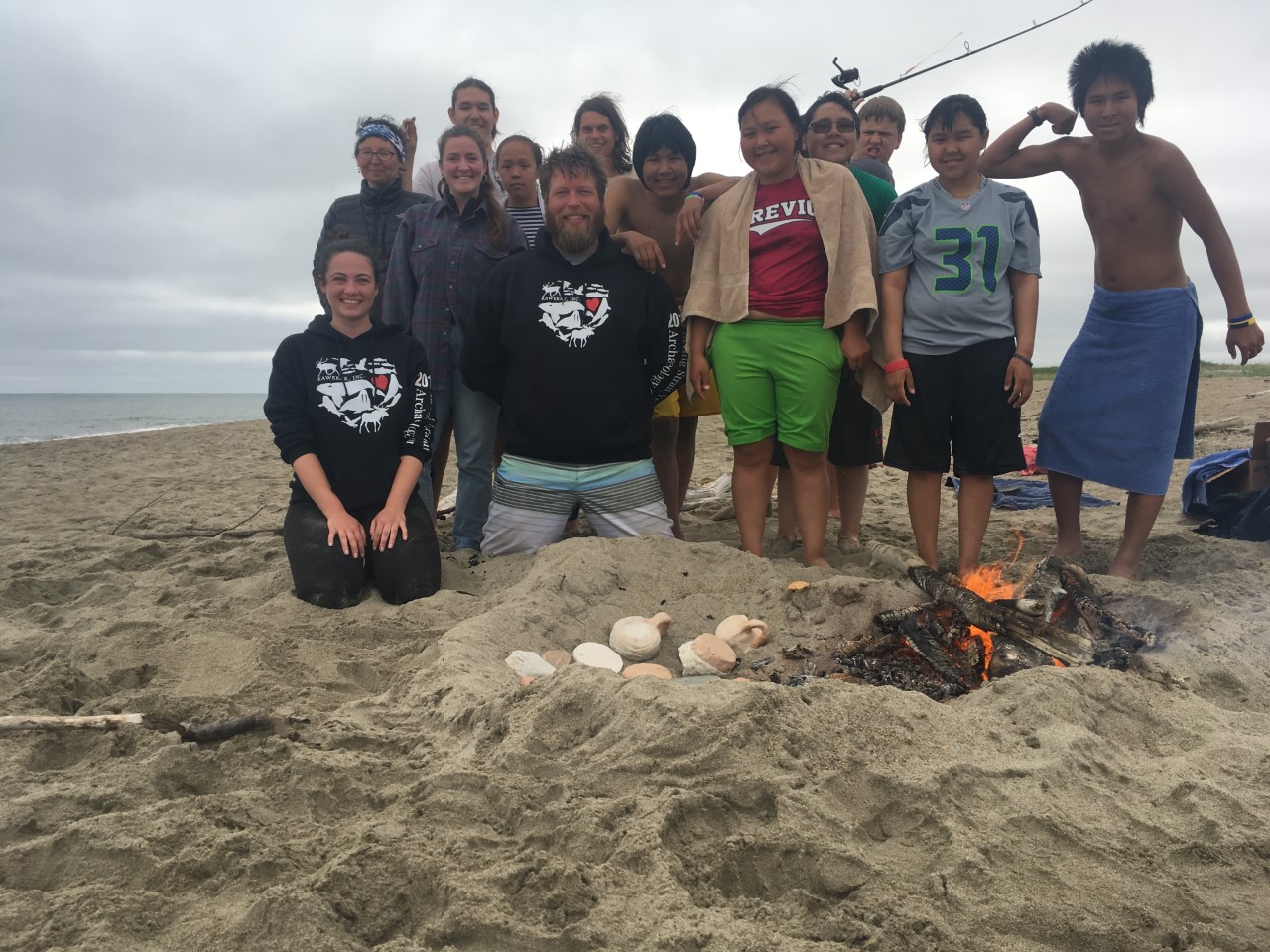 A group of adults and kids smile for a photograph on the beach with a fire in view.
