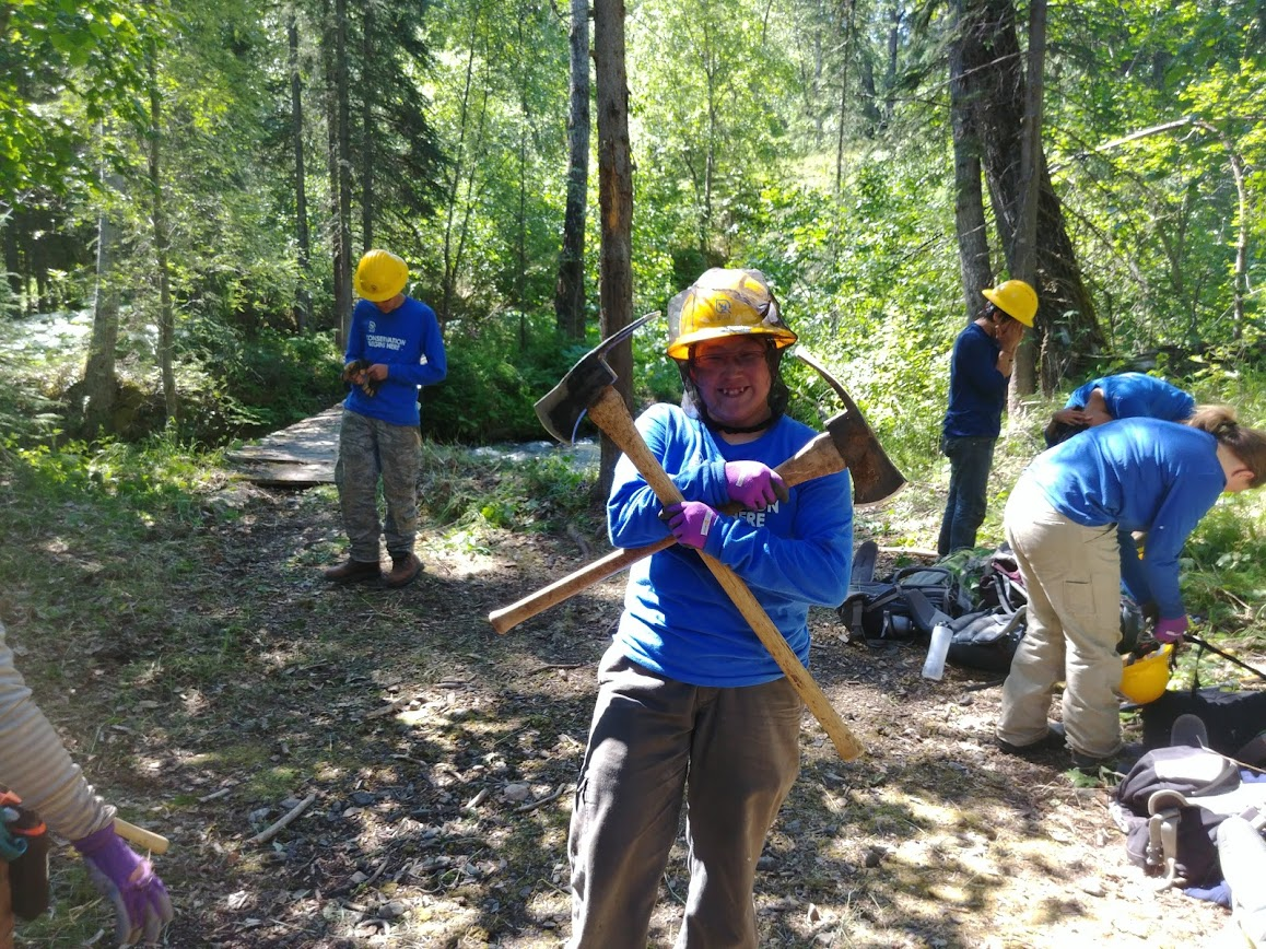 a trail crew member wearing a hard hat holds two pulaskis in a cross formation