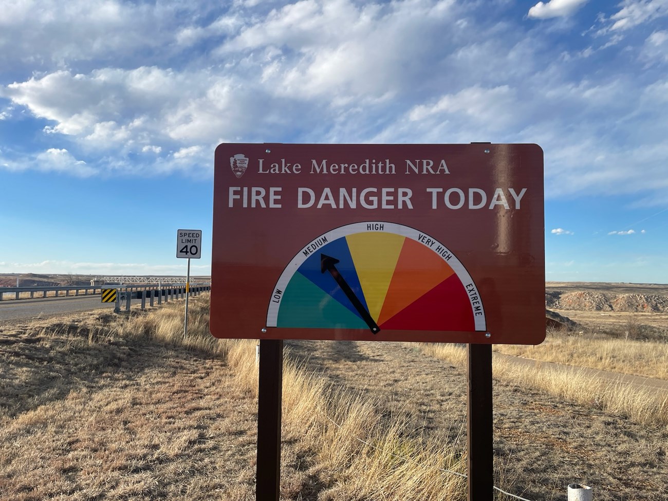 Brown sign, text reads: "Lake Meredith NRA Fire Danger Today." A green, blue, yellow, orange, and red semicircle with an arrow pointing to the blue color reads, "low, medium, high, very high, extreme."