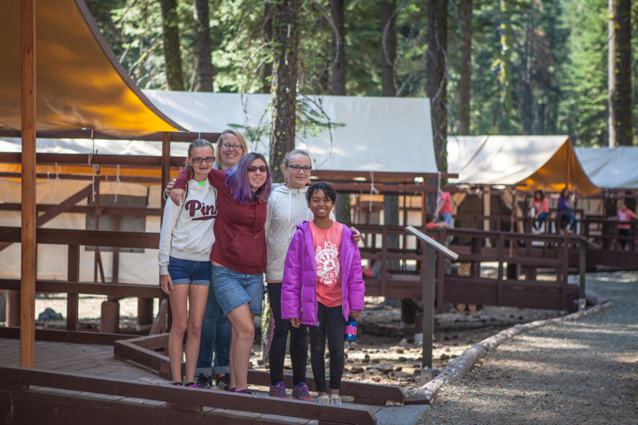 Five girls standing close pose for a photo on a wooden walkway outside of canvas tent at a youth camping facility