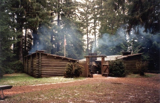 A fort made of logs stands in a forest clearing. The fort is made of two buildings, connected by a tall log fence and gate. There is smoke rising from the building’s chimneys.