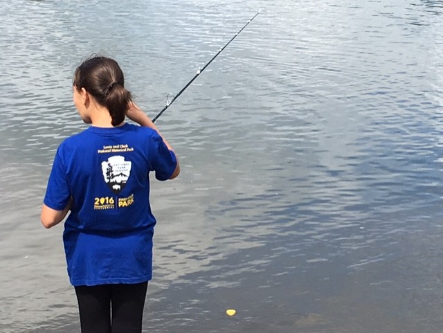 A woman in a blue national park shirt stands at the edge of a river with a fishing pole in hand.