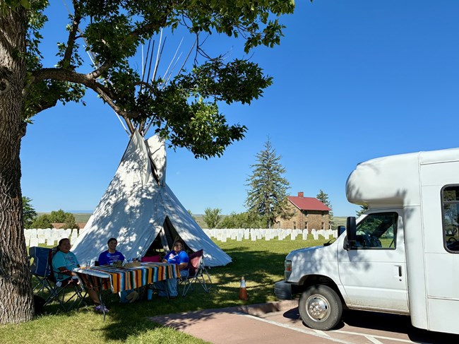 Three people sit in front of a tipi.
