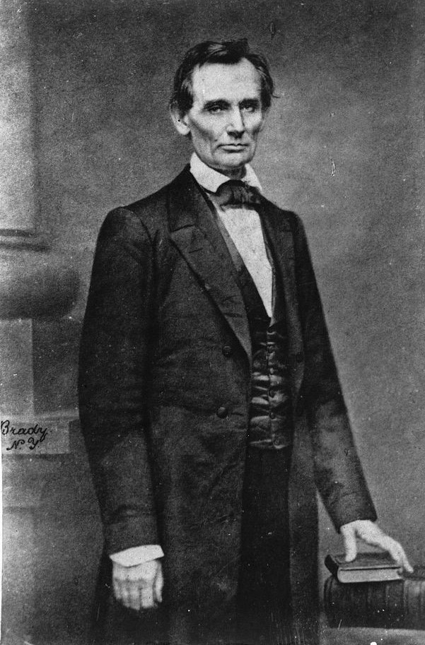 Clean shaven Abraham Lincoln in suit standing with hand resting on table.
