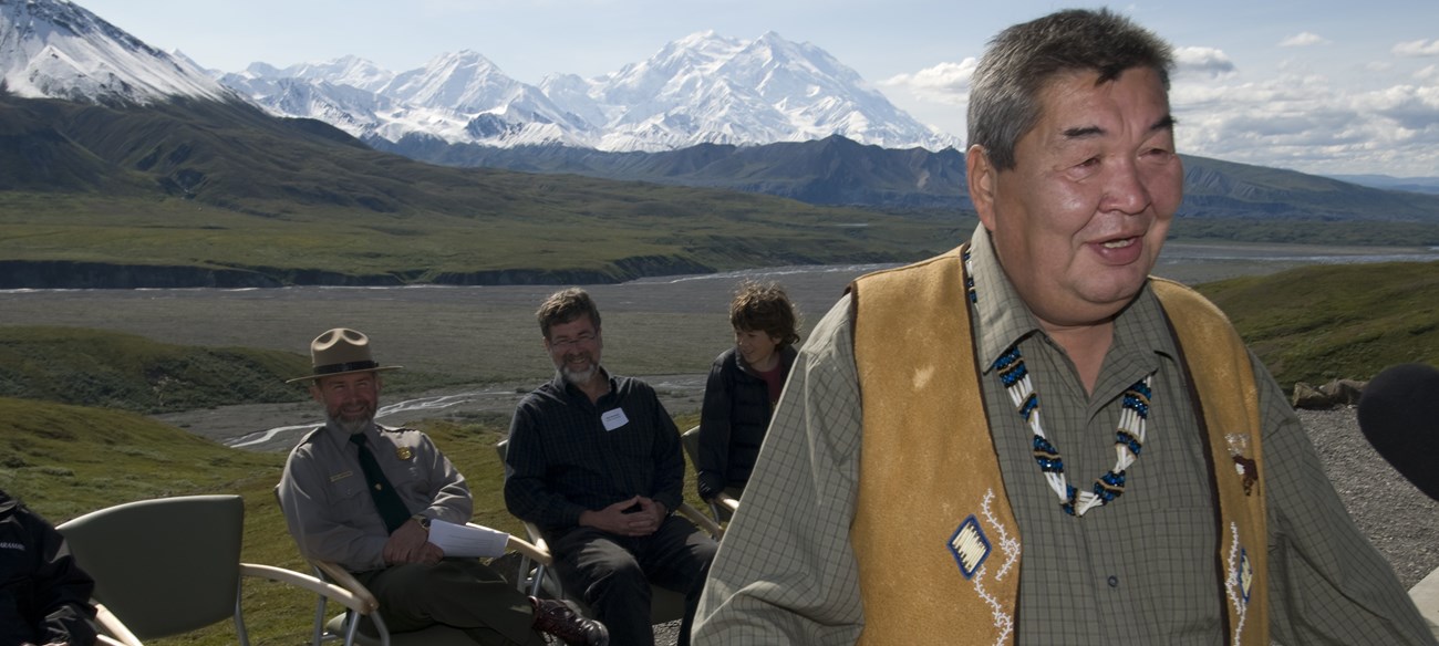 Chief Mitch Demientieff speaks at a podium in Denali National Park and Preserve with park staff and mountains in the background