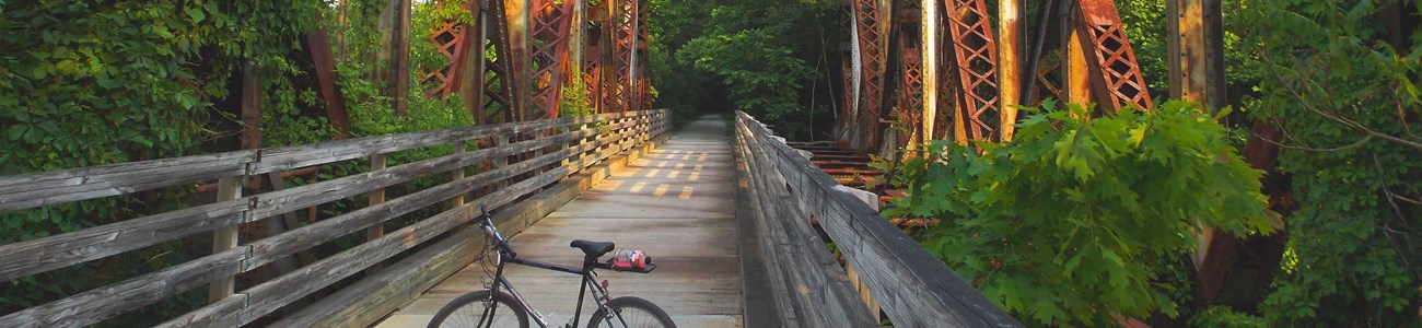 A bicycle stands across a boardwalk trail built into a historic bridge, seen overhead.