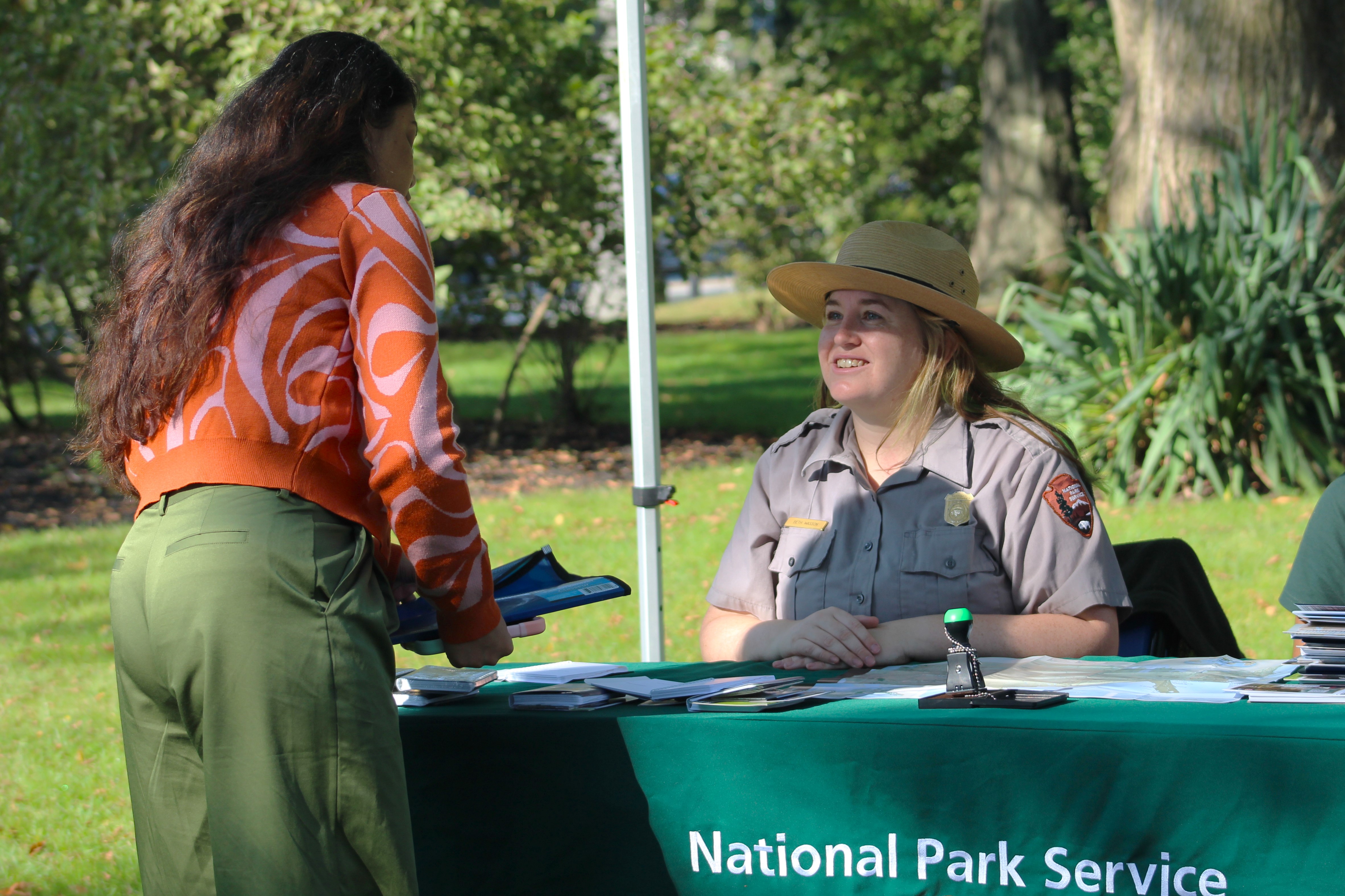 Ranger seated under a "National Park Service" outdoor tent speaking with visitor