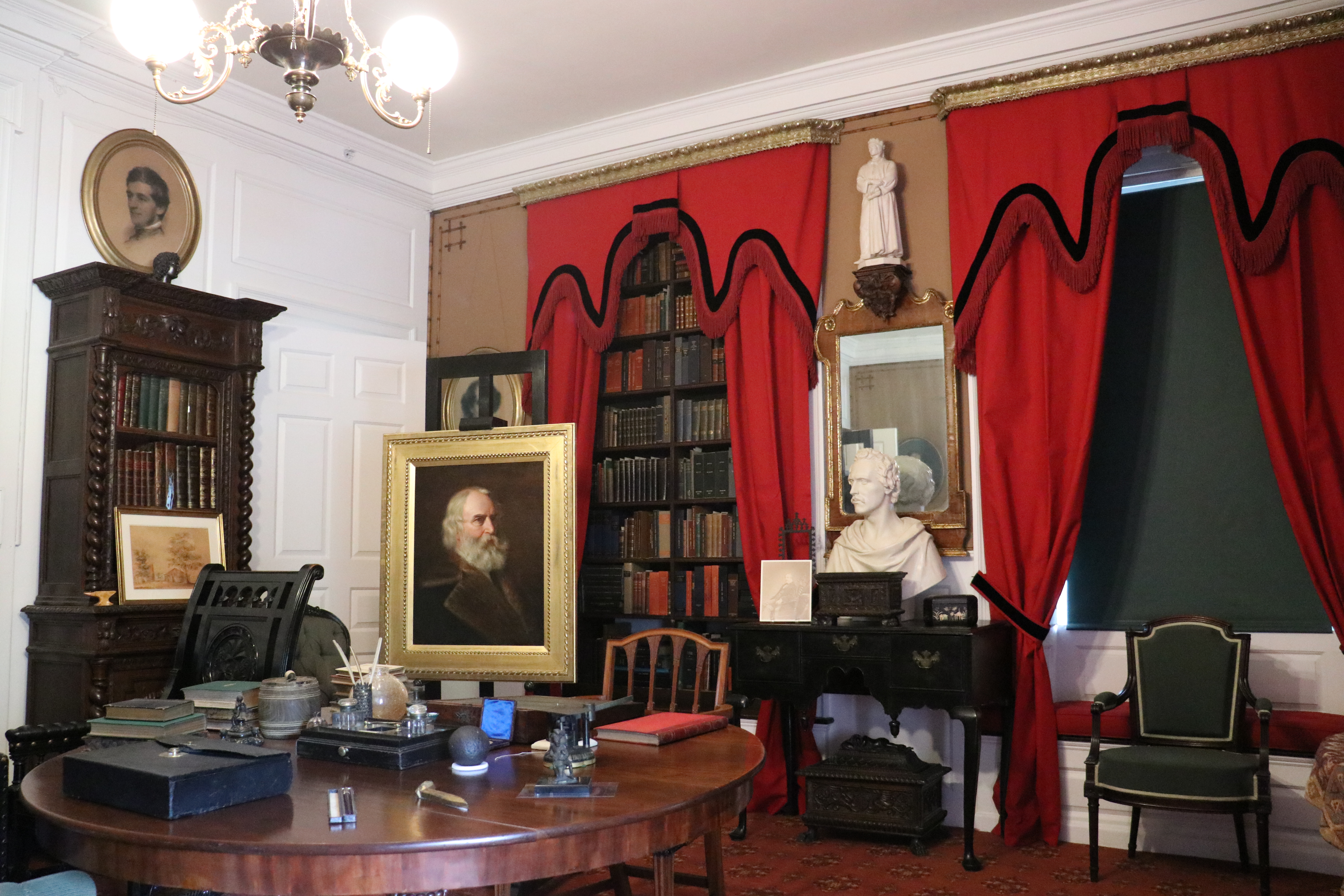 View of historic furnished room with round center table with books and inkwells, red curtains with black trim, and oil portrait of Henry Longfellow on easel.