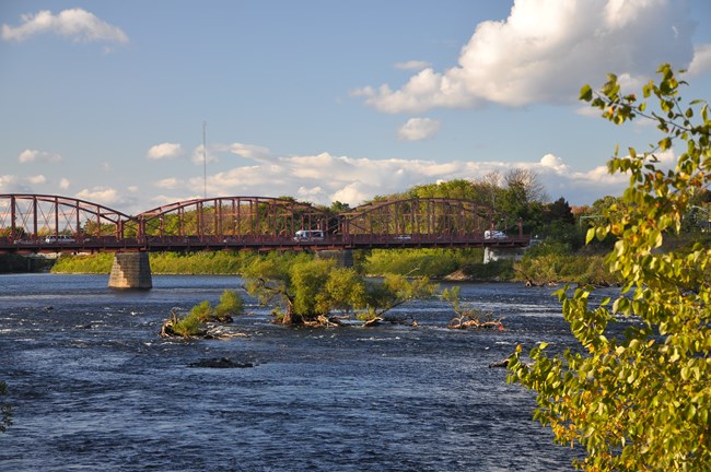 A large arched bridge sits in the background suspended above the merrimack River, with small islands populated with autumnal trees in the foreground and middle of the view
