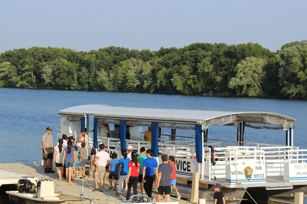 A group of people walk along a dock leading to a riverboat where a ranger waits