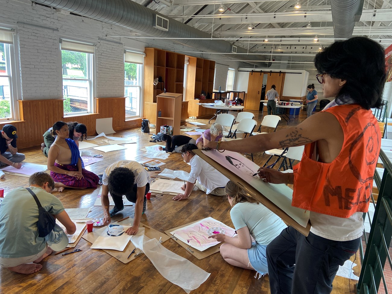 A group of people kneeling, sketching and painting on paper with a dancer posed among them, one artist stands with a orange vest sketching the dancer.