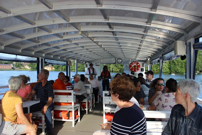 A group of people sit on benches on a boat while a person wearing a life jacket speaks into a microphone