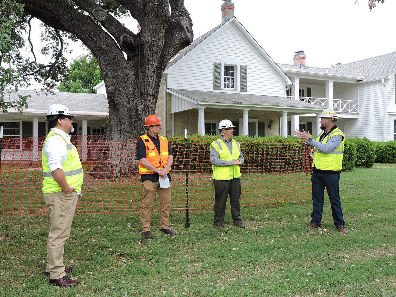 Four people in bright safety vests and hard hats stand in front of a two-story house surrounded by orange fencing.