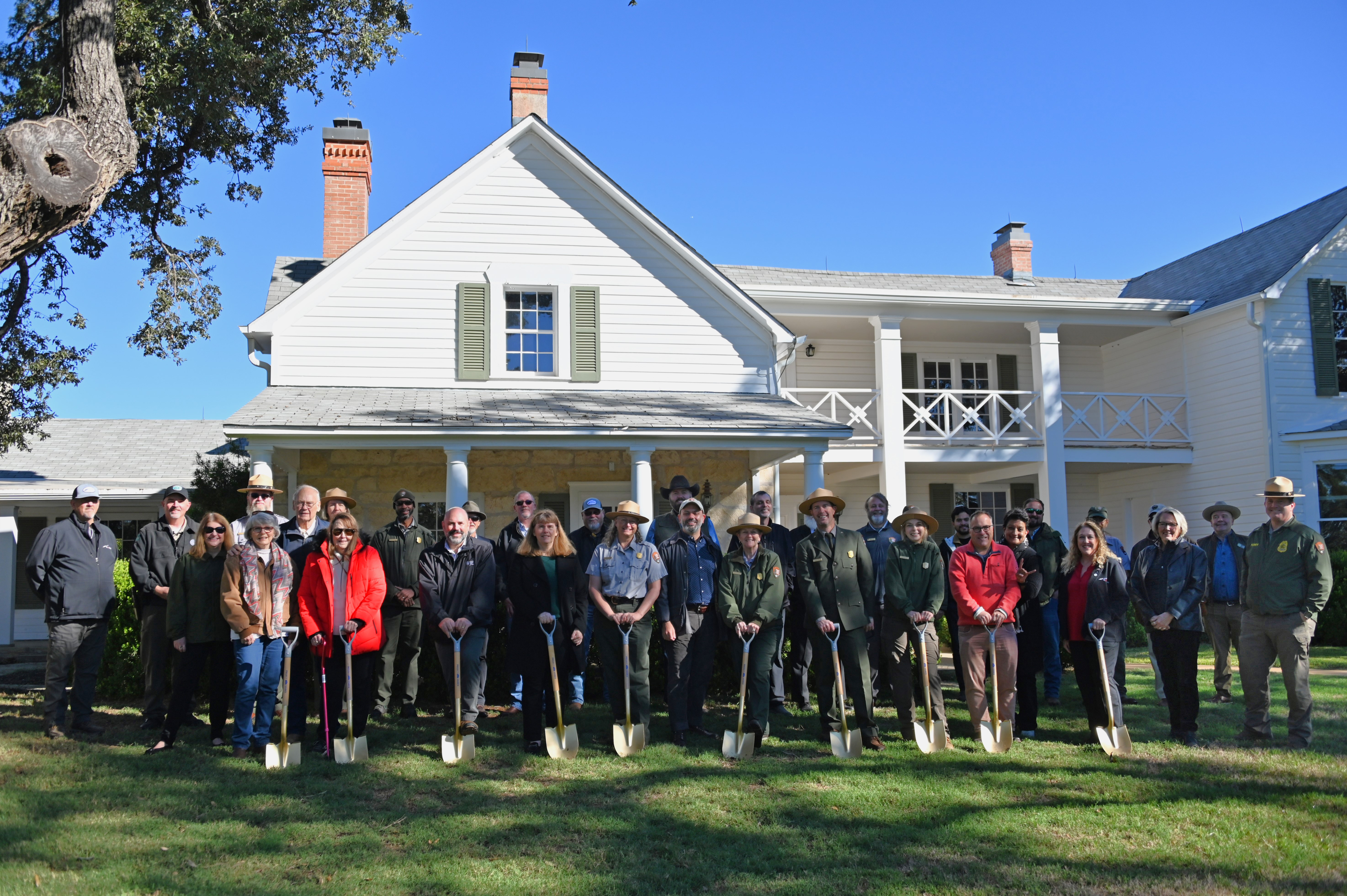 A large group of people, some holding gold shovels, stand in front of a big, two-story, white frame house.