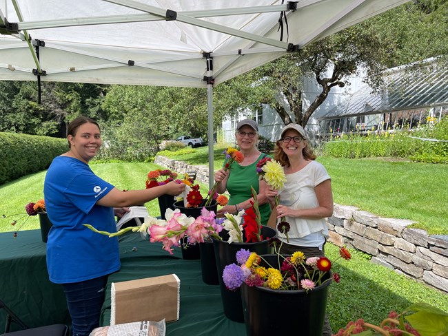 staff member in blue shirt and two park visitors smile at camera holding flower bouquets