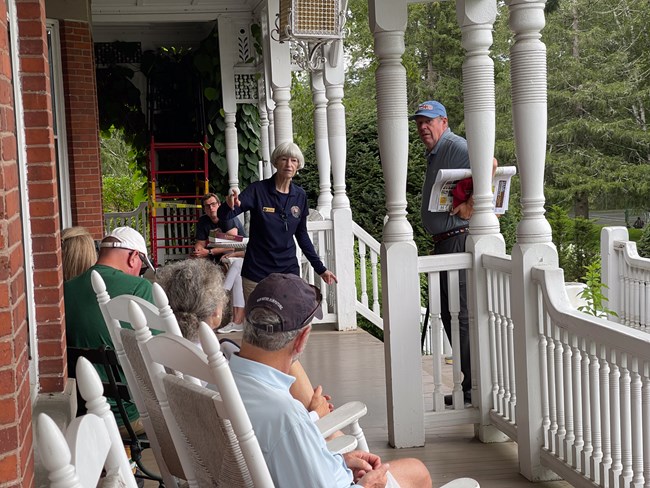 park visitors sit on mansion porch while park volunteer talks