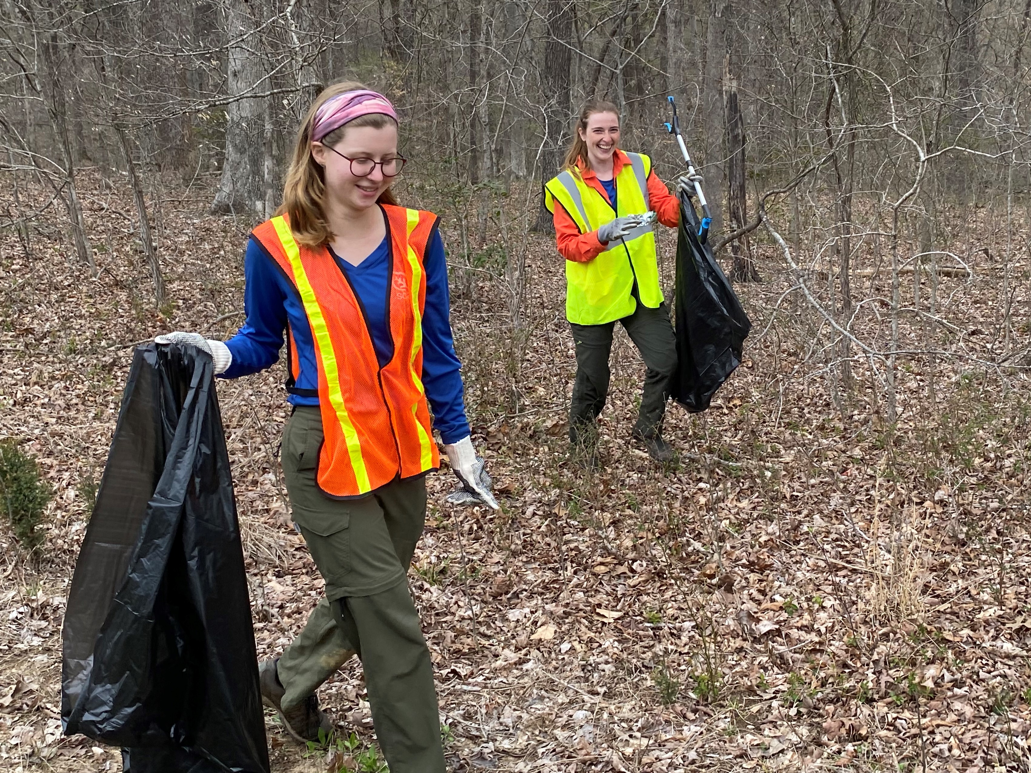 Two people wearing orange and yellow safety vests carry trash bags and while walking in a wooded area.