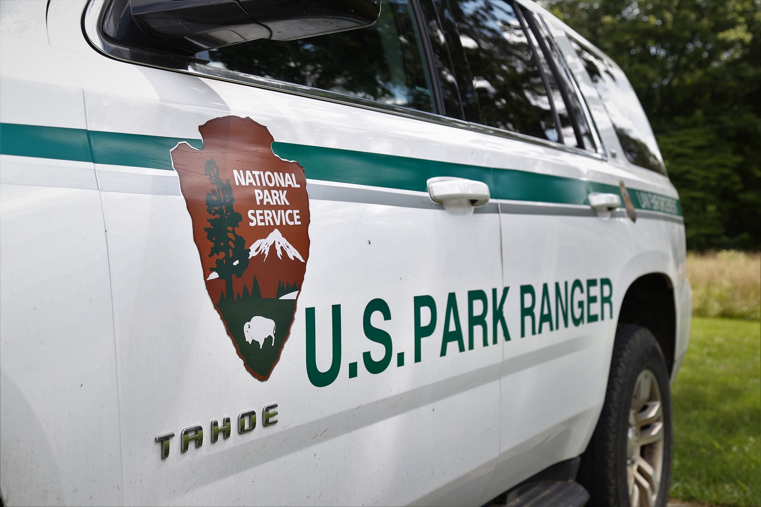 The side of a white vehicle with the brown, green and white National Park Service Arrowhead Logo and the words "U.S. Park Ranger" written in green.