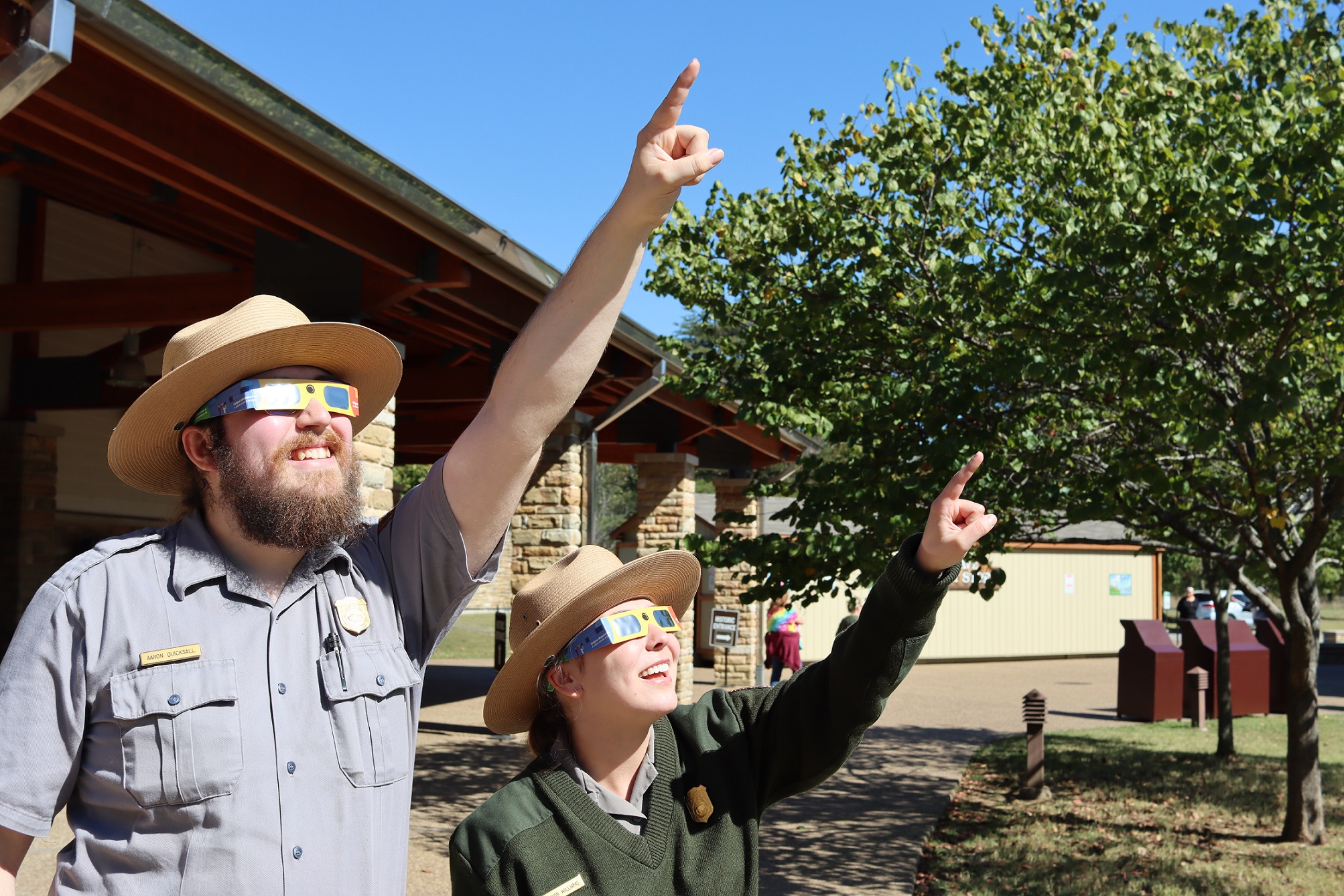 Two people wearing National Park Service uniforms and colorful dark lensed glasses point toward a sunny sky.