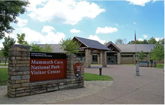 A stone sign with NPS arrowhead and white text "Mammoth Cave National Park Visitor Center" with a large stone building in the background.