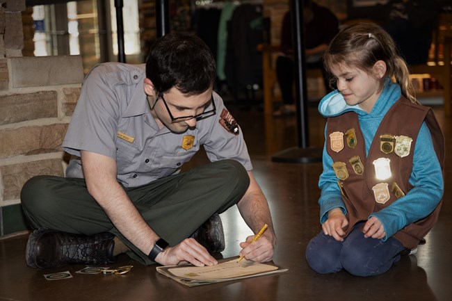 A person in a park ranger uniform signs his name on a Junior Ranger certificate for the child sitting next to him.