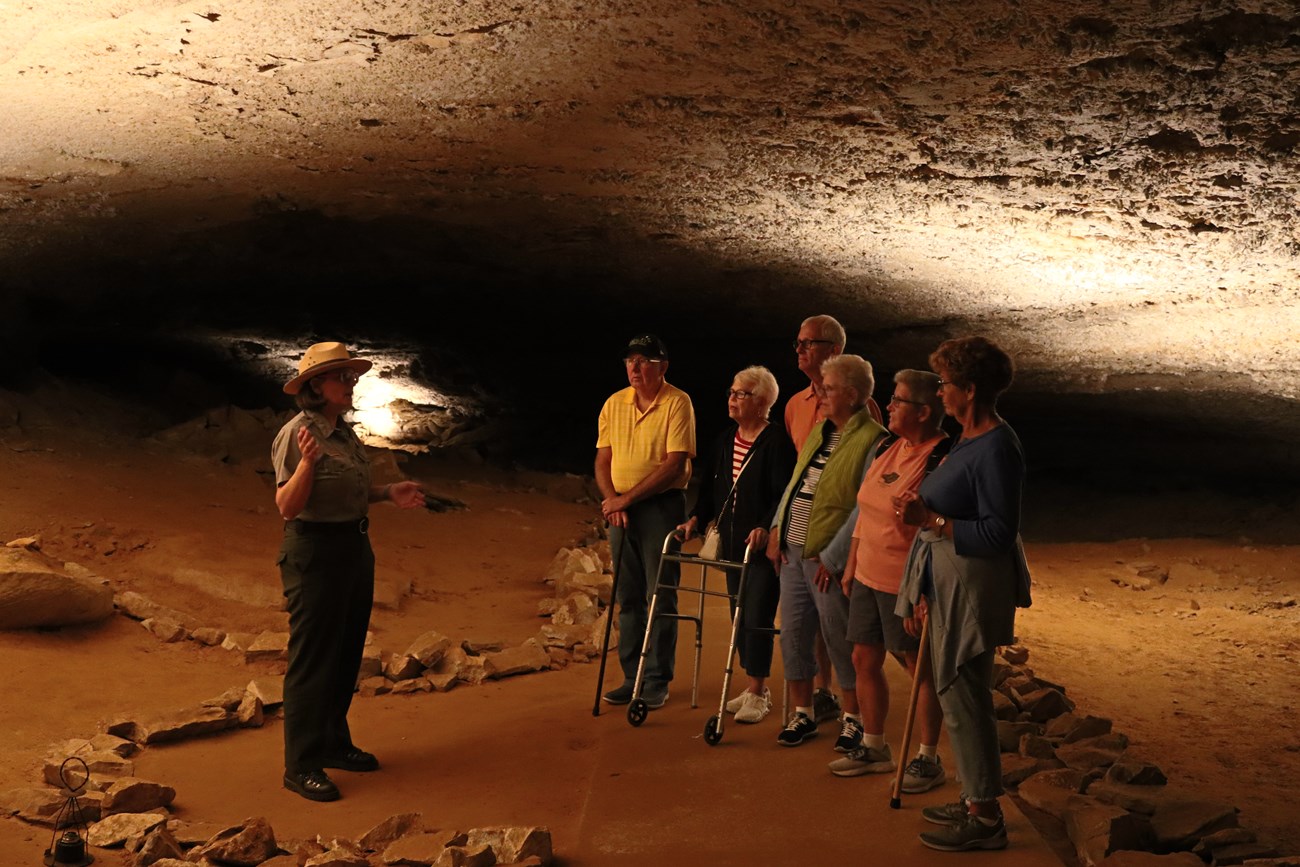 An NPS ranger addresses a group of 6 people, some using assistive devices like canes or a walker, in an underground passageway.