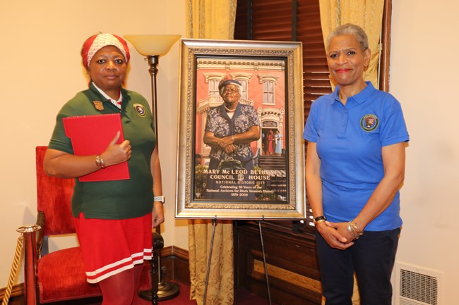 Two volunteers stand in front of a framed portrait