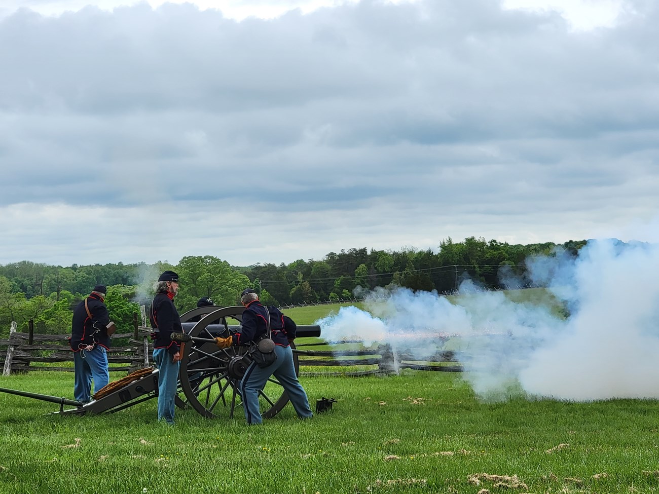The Bull Run Legion fire's the park's cannon wearing Federal uniforms in blue with red trim. Smoke and fire pour out of the barrel against a green, luch backdrop.