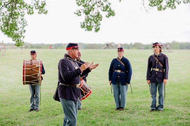 People in blue and red uniforms in the Ft. McHenry Fife and Drum Corps demonstrate period music. There is a fifer in the foreground and 4 people in the background- 2 with drums, 2 with fifes.