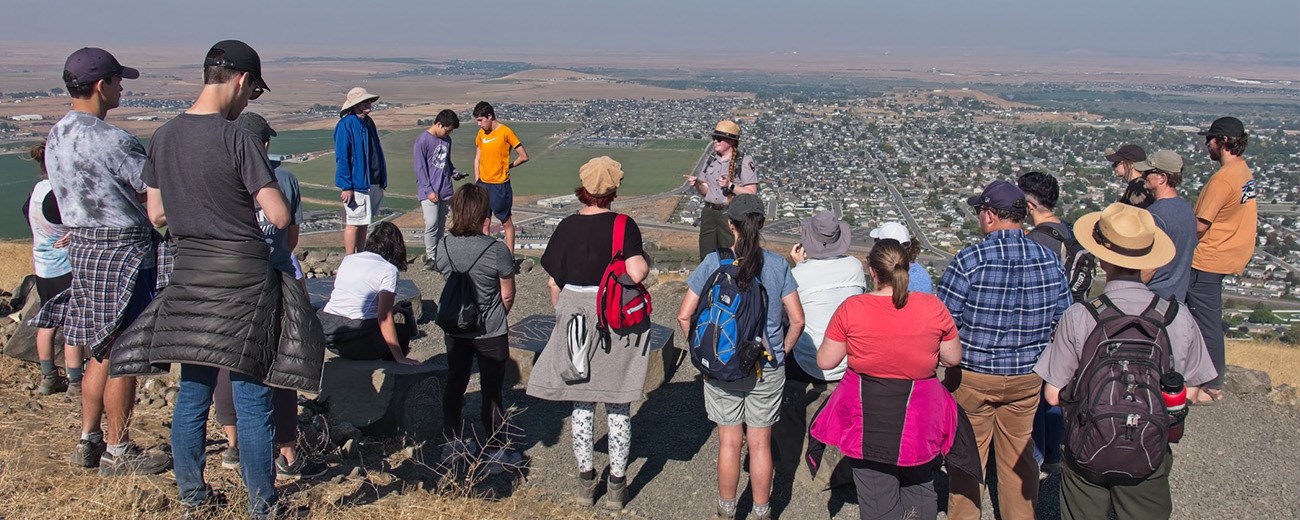 A woman in a ranger hat talks to a large group atop a big hill overlooking a city.