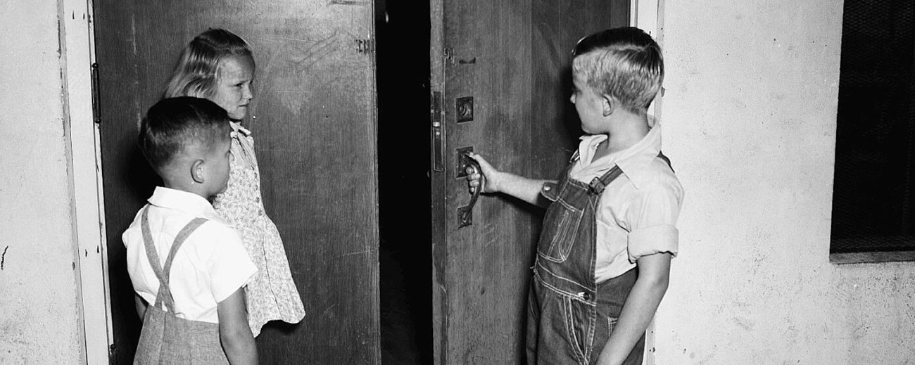 3 elementary students walk enter a room with one boy holding open the door.