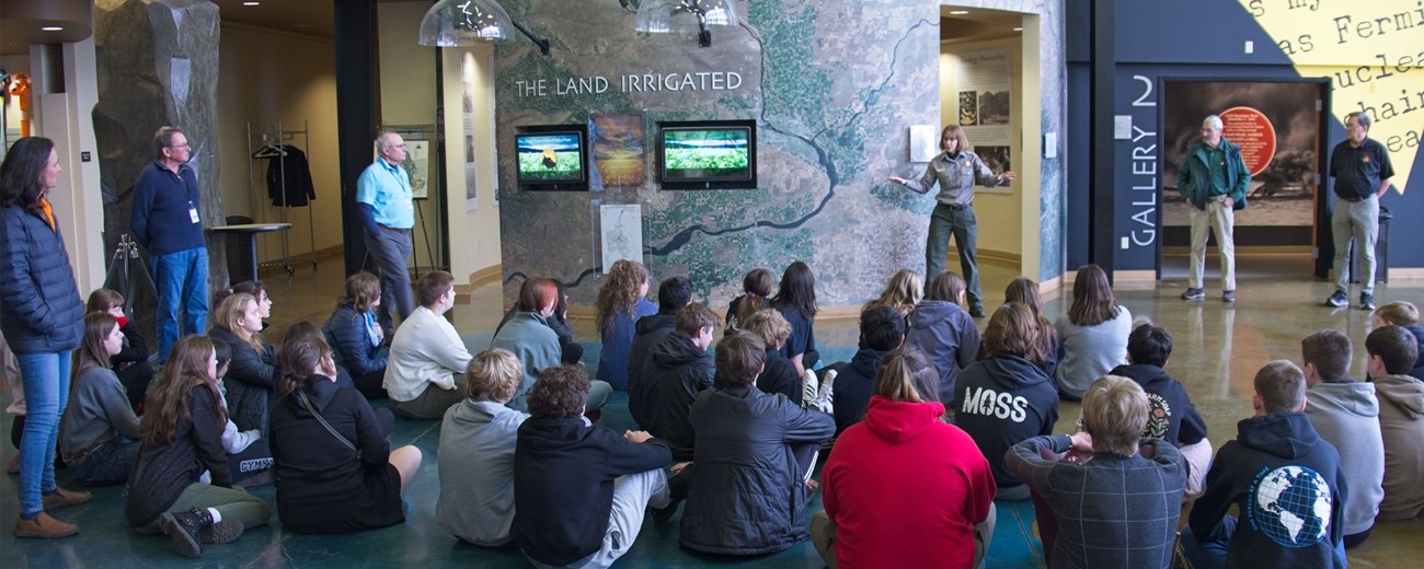 35 students sit on the floor in an open room and 5 adults stand looking at a uniformed ranger talking